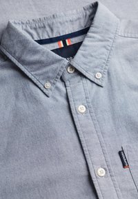 Lichtblauw overhemd met knoopsluiting, witte knopen en een klein "Superdry" stoffen label op het borstzakje.