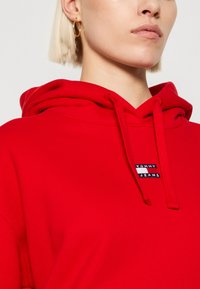 Raudona gobtuvė pagaminta iš minkštos medžiagos, su priekiu kangaroo tipo kišene, raišteliu ant gobtuvo ir maža Tommy Jeans logotipo etikete ant krūtinės.