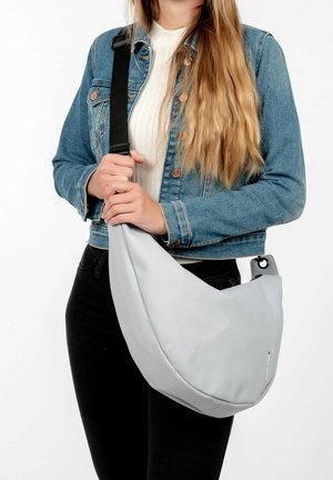 Personne portant une veste en jean et un haut blanc, portant un grand sac à bandoulière gris clair avec une sangle noire en travers du torse.