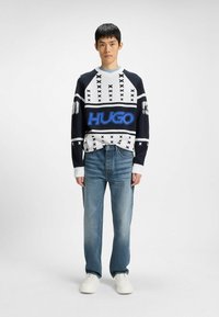 Maglione blu e bianco con un motivo geometrico, caratterizzato da un audace logo blu "HUGO"; abbinato a jeans azzurri e sneakers bianche.