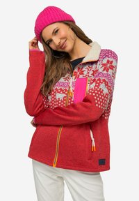 Giacca in pile rossa con motivi fair isle, inclusi animali, e una tasca con zip rosa. Presenta un colletto in pelliccia sintetica bianca e un orlo con cordoncino.