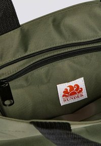 Interno di una borsa di tessuto verde con una cerniera nera, materiale testurizzato e un'etichetta bianca con un logo del sole rosso di Sundek.