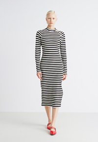 Črtasta dolga obleka s dolgimi rokavi v črni in beli barvi, prilegajoča silhueta, do kolen z okroglim izrezom, vsebuje dekorativni dodatek blizu rame.