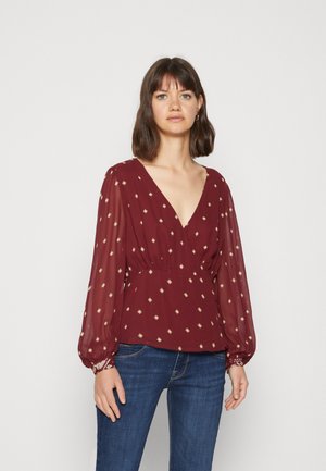 Blouse bordeaux avec un décolleté en V profond, des manches longues bouffantes et des motifs en diamants dorés, fabriquée en tissu léger, assortie à un jean bleu foncé.