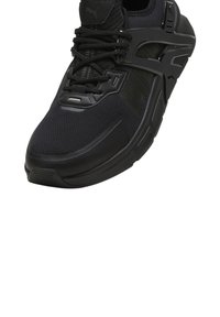 Scarpa sportiva nera con tomaia in rete, suola in gomma, design testurizzato e sistema di allacciatura evidente. Presenta accenti di branding pronunciati.