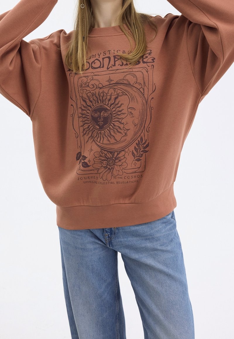 Světle hnědá mikina s volným střihem, zdobená grafikou slunce a měsíce v tmavě hnědé barvě, s textem níže popisujícím kosmickou cestu.