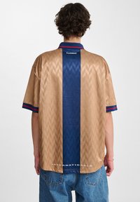 Kortærmet trøje i beige med et tekstureret diamantmønster, med en blå lodret stribe, blå krave og teksten "INTERNATIONALS".