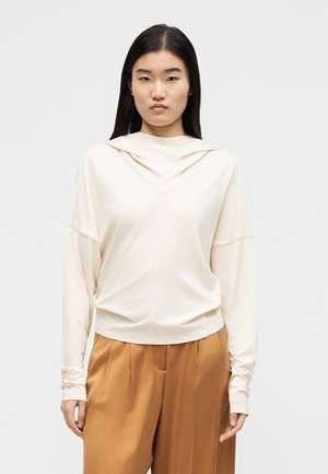 Žena s dlouhými tmavými vlasy, oblečená v volném krémovém tričku s dlouhým rukávem a vysokým pasem šírokých béžových kalhotách, stojící před neutrálním pozadím.