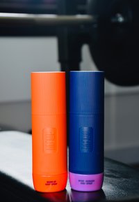 Zwei zylindrische Körpersprayflaschen, eine orange mit gerippter Textur, beschriftet mit "Warm-Up", die andere blau mit pinkem Boden, beschriftet mit "Sport Recovery."