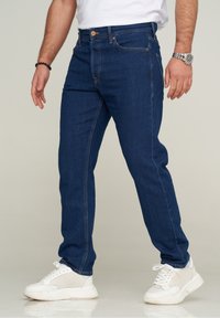 Man som bär mörkblå jeans, vita mesh-sneakers, en vit t-shirt, ett svart pärlarmband och ett silverfärgat armbandsur som står framför en ljus bakgrund.