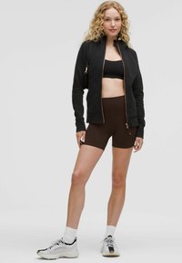 Black zip-up jacket with a high collar, brown high-waisted shorts, and white athletic sneakers. Textured fabric and fitted design.