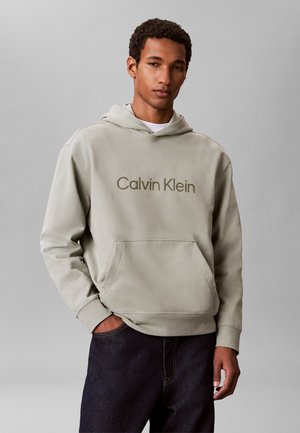 Mand iført en lysegrå Calvin Klein hættetrøje og mørke jeans, stående med den ene hånd i lommen mod en ensfarvet grå baggrund.