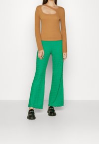 Ruskea ribattuja pitkähihainen paita, jossa on leikattu olkain yksityiskohta, yhdistettynä tekstuuroituihin vihreisiin leveisiin housuihin ja mustiin paksupohjaisiin mokkasiineihin.