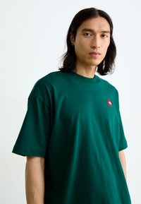 Tricou verde din bumbac, cu guler rotund și mâneci scurte, având un mic logo roșu pe partea stângă a pieptului. Textură moale, croială lejeră.