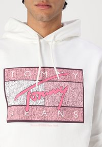 Hvit hettegenser med et flerfarget grafikk der "TOMMY" er i rosa og "JEANS" i hvitt mot teksturerte striper. Snøring ved halsen.