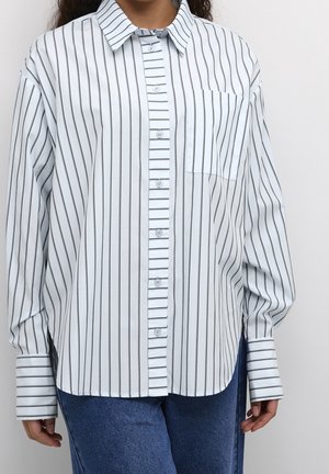 Personne portant une chemise blanche à manches longues avec de fines rayures verticales noires et un jean bleu sur un fond uni.