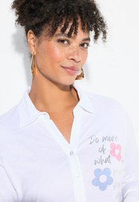 Weißes Hemd mit Kragen, das bedruckten Text und florale Designs in Pink und Blau aufweist, mit einer glatten Stofftextur und glänzenden Knopfdetails.