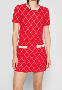 Mujer vestida con una blusa roja de manga corta y una falda a juego con un patrón de diamantes y pequeños adornos similares a perlas.