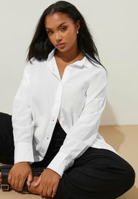 Witte langemouw blouse van lichtgewicht stof met een relaxte pasvorm. Voorzien van een kraag en knoopdetail, gedragen over zwarte broek.