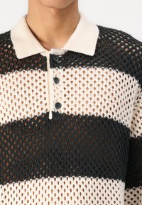 Polo en tricot avec un design en maille, présentant des rayures alternées noir et crème, un col crème et trois boutons noirs au plastron.