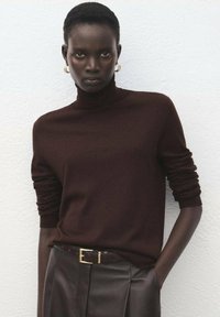 Pulover maro cu guler înalt, realizat din material moale, prevăzut cu mâneci lungi. Asociat cu pantaloni maro din piele cu talie înaltă și o curea cu accente aurii.