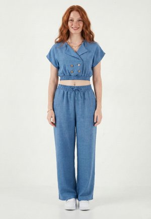 Jeune femme aux longs cheveux roux portant un haut bleu court à double boutonnage et un pantalon large assorti, debout et souriante.