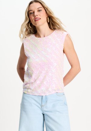 Vrouw met lang blond haar draagt een mouwloze roze paillettetop en lichtblauwe spijkerbroek, staand met de handen achter de rug tegen een witte achtergrond.