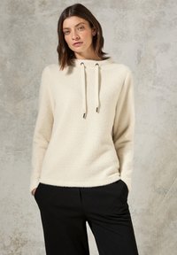 Pullover en polaire couleur crème avec une capuche, pr�ésentant une surface texturée, des cordons de serrage réglables et une coupe décontractée, associé à un pantalon noir.