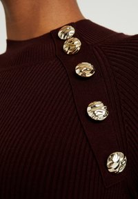 Pull en maille côtelée bourgogne avec quatre boutons dorés brillants et texturés sur l'épaule, doté d'un col rond haut.