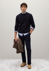 Maglione navy su una camicia a righe blu chiaro, pantaloni scuri, tiene un blazer a scacchi marrone e indossa scarpe in suede color sabbia. Sfondo semplice.