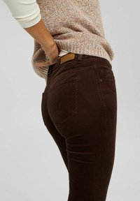 Pantalon en velours côtelé marron à coupe ajustée, avec poches arrière et patch en cuir sur la ceinture. La texture est côtelée et douce.