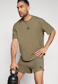 Αθλητικό σετ ελιάς πράσινου - σορτς και μπλούζα, και τα δύο με μαύρο λογότυπο adidas. Το υλικό φαίνεται υφασμάτινο. Το μοντέλο κρατάει μια μαύρη kettlebell.