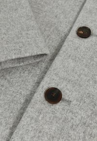 Hallhalli villakangas, millel on klapp ja ümmargune tumepruun nupp nelja auguga, millel on pehme tekstuur.