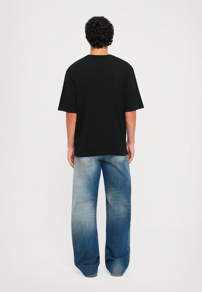 Uomo in piedi di spalle che indossa una maglietta nera larga e jeans a gamba larga blu sbiaditi, davanti a uno sfondo bianco semplice.