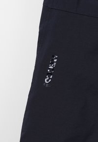 Marineblauwe stoffen broek met een logo in reflecterend materiaal op de linkerdij, met een gladde textuur en functioneel ontwerp.