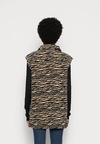 Fleeceväst i beige med svarta tigerrandiga mönster, med hög krage och ärmlös design, i kombination med en svart långärmad skjorta.