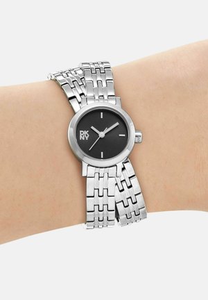 Montre-bracelet à maillons argentés avec cadran rond noir, marqueurs minimalistes et logo DKNY, portée sur un poignet à peau claire.
