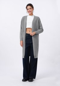 Grijze lange cardigan van zacht materiaal, met een open voorkant, geribbelde manchetten en twee voorzakken, gecombineerd met donker wijde jeans.