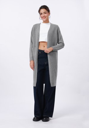 Grijze lange cardigan van zacht materiaal, met een open voorkant, geribbelde manchetten en twee voorzakken, gecombineerd met donker wijde jeans.