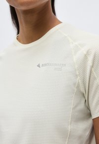 Κοντινό πλάνο ενός ατόμου που φοράει ανοιχτό μπεζ κοντομάνικο πουκάμισο με μικρό πουά σχέδιο και αχνό λογότυπο στο στήθος.