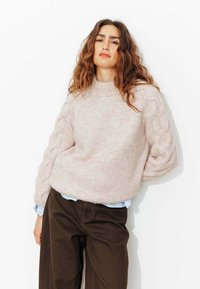 Pull en tricot rose clair avec un col montant et des détails en tricot torsadé sur les manches, associé à un pantalon marron foncé et une chemise bleue en dessous.