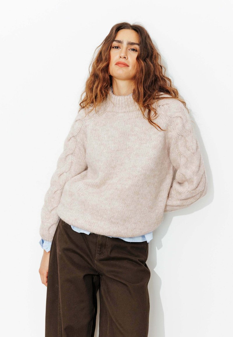 Pull en tricot rose clair avec un col montant et des détails en tricot torsadé sur les manches, associé à un pantalon marron foncé et une chemise bleue en dessous.