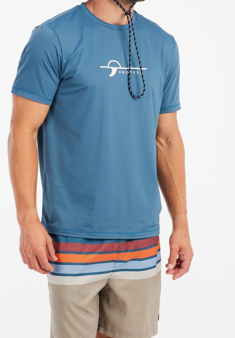 Homme portant une chemise bleue à manches courtes avec le logo « Protest » et un short de bain beige avec des rayures horizontales rouges, oranges, bleues et blanches.