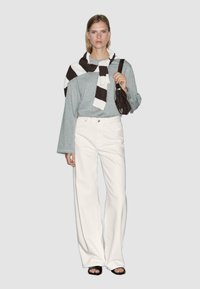 Maglia grigia a maniche lunghe, jeans bianchi a gamba larga, sciarpa a righe e una borsa scura. La modella indossa sandali neri con tacco; sfondo neutro.