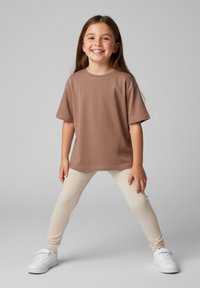 Brązowy T-shirt z krótkim rękawem w połączeniu z beżowymi legginsami i białymi tenisówkami. Koszulka ma zaokrąglony dekolt i luźny krój.