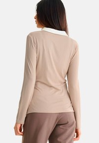 Beige langwerpige top met verticale ribbels, voorzien van een witte kraag. De stof lijkt glad met een getailleerde vorm, gedragen met een bruine broek.