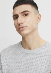 Pulover tricotat de culoare gri deschis, cu un model texturat, gât rotund cu marginile ribate și manșete ajustate. Prim-plan al unui model care prezintă articolul vestimentar.