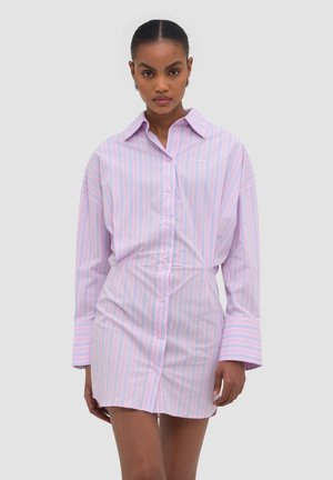 Kobieta ubrana w różowo-niebieską, cienko paskowaną sukienkę koszulową z długim rękawem i szerokimi mankietami, stojąca na jednolitym tle.