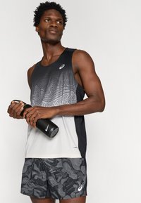 Ανδρικό αθλητικό αμάνικο μπλουζάκι σε μαύρο και γκρί με σχέδιο κυμάτων; ελαφρύ ύφασμα. Συνδυασμένο με κοντά σορτς με σχέδιο. Κρατάει μια μαύρη μπουκάλι νερού.