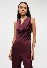 Macacão sem mangas bordeaux com decote drapeado, textura de cetim suave e cintura ajustada. Apresenta costuras mínimas e nenhum hardware visível.